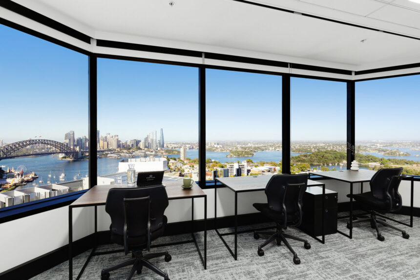 Three minimalist office desks paired with black ergonomic chairs face expansive floor-to-ceiling corner windows, offering a scenic high-rise panoramic view of Sydney Harbour and the city skyline.