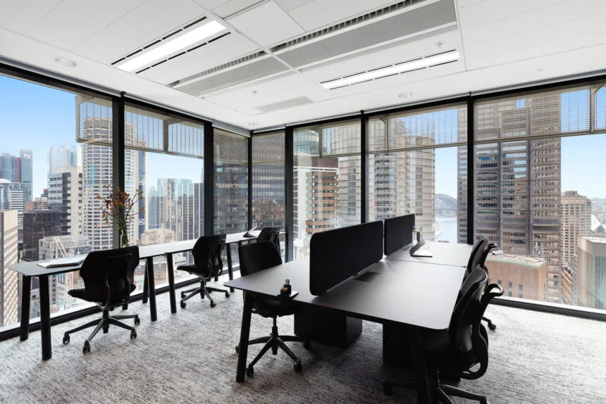Black office desks with ergonomic chairs are arranged inside a modern corner office, surrounded by floor-to-ceiling windows that overlook a sunlit, high-rise city skyline.