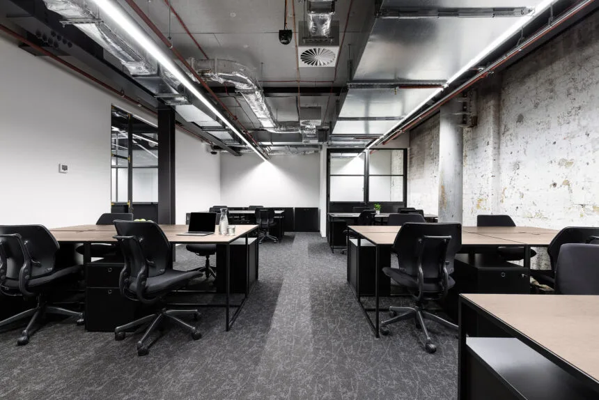 Modern wooden desks paired with black ergonomic chairs fill an open office featuring dark patterned carpet, exposed ceiling ductwork with linear strip lights, and a distressed white brick wall.