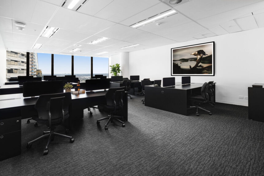 Black office desks and ergonomic chairs sit arranged inside a spacious, modern workspace featuring dark gray carpeting, large windows offering city views, and framed artwork hanging on a white wall.