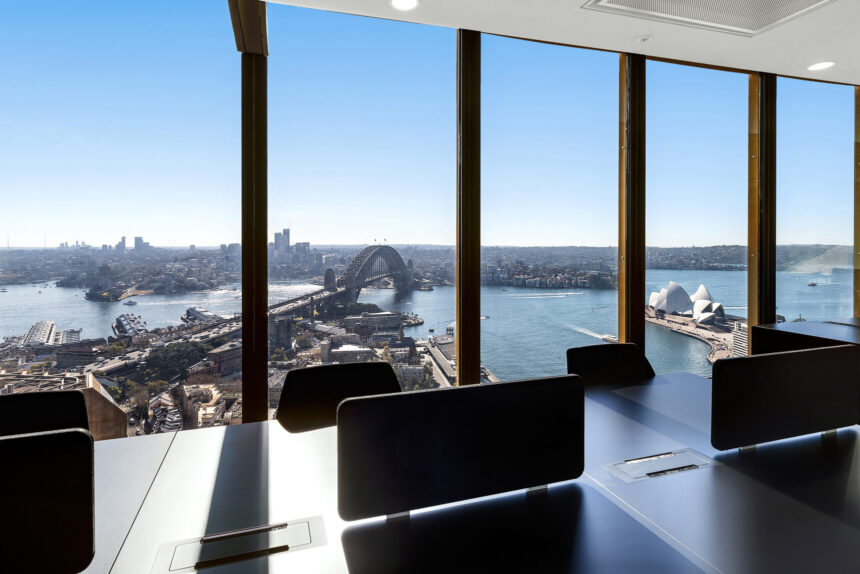 Dark office desks with privacy screens sit before floor-to-ceiling windows, offering a panoramic high-rise view of the Sydney Harbour Bridge and Opera House under a clear blue sky.