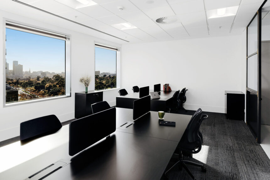 Black office desks paired with ergonomic chairs sit neatly arranged inside a bright, modern office space featuring large windows with scenic views of a city skyline and parkland.