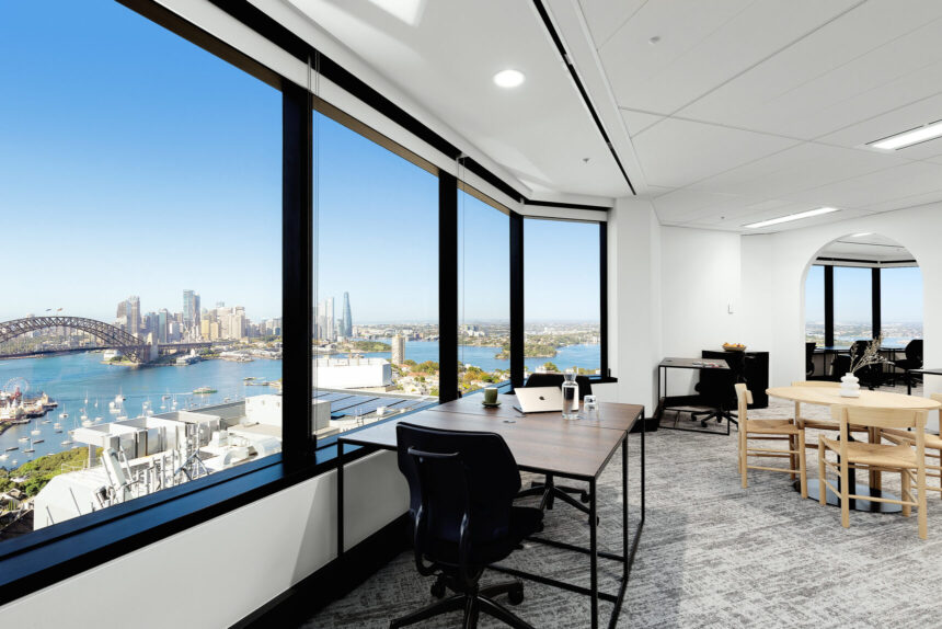 A wooden office desk sits beside expansive floor-to-ceiling windows inside a modern high-rise workspace, offering panoramic views of a sunny city harbour and bridge.