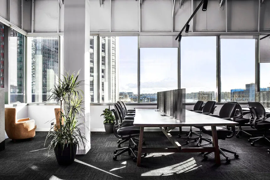 A long wooden desk lined with black office chairs fills a modern open office, framed by potted plants and large floor-to-ceiling windows overlooking a bright city skyline.
