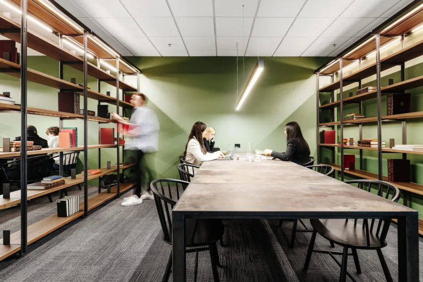 People sit and work at a long conference table while another person browses tall bookshelves in a modern, green-walled office library.
