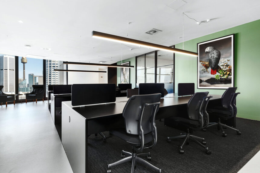Modern black desks with ergonomic chairs line a sleek, open-plan office space featuring a green accent wall with framed art, linear hanging lights, and floor-to-ceiling city views.