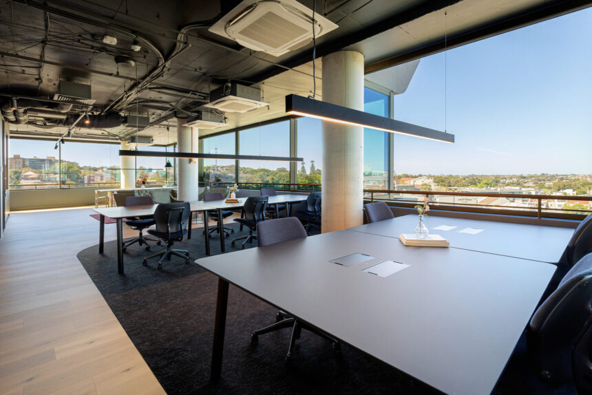 Dark modern office desks and ergonomic chairs stand inside a bright, open-plan workspace beneath hanging linear lights, with floor-to-ceiling windows overlooking an expansive city landscape.