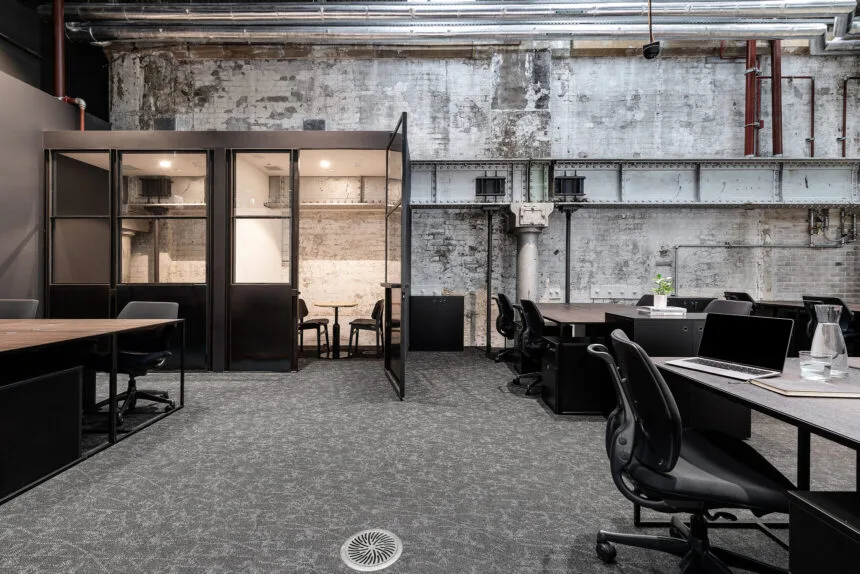 Dark wooden desks and ergonomic chairs furnish an open industrial office featuring glass meeting pods, set against weathered brick walls with exposed pipes and structural steel beams.