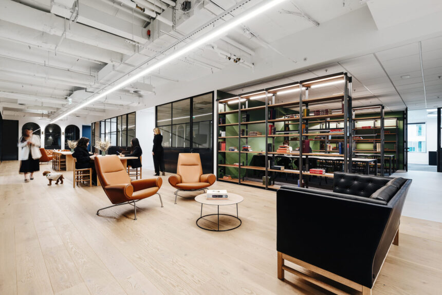 Two cognac leather armchairs and a black sofa surround a coffee table while people work and walk in a spacious, modern office featuring industrial ceiling fixtures and metal shelving.