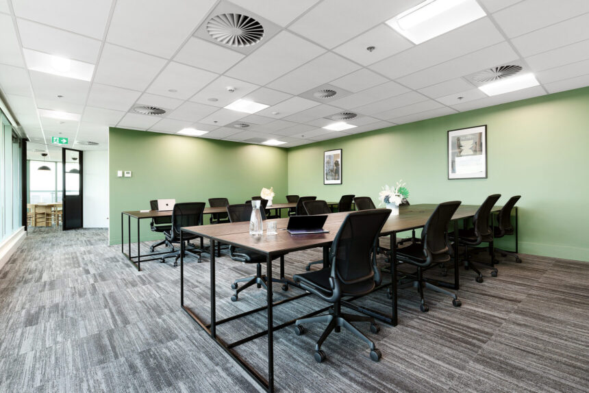 Black office chairs surround long dark-wood desks equipped with laptops inside a modern office room featuring sage-green walls, grey patterned carpet, and recessed ceiling lights.
