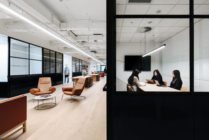 Three people work around a table inside a glass-framed meeting room, overlooking a spacious, modern office corridor featuring brown leather armchairs, light wooden floors, and exposed ceiling pipes.