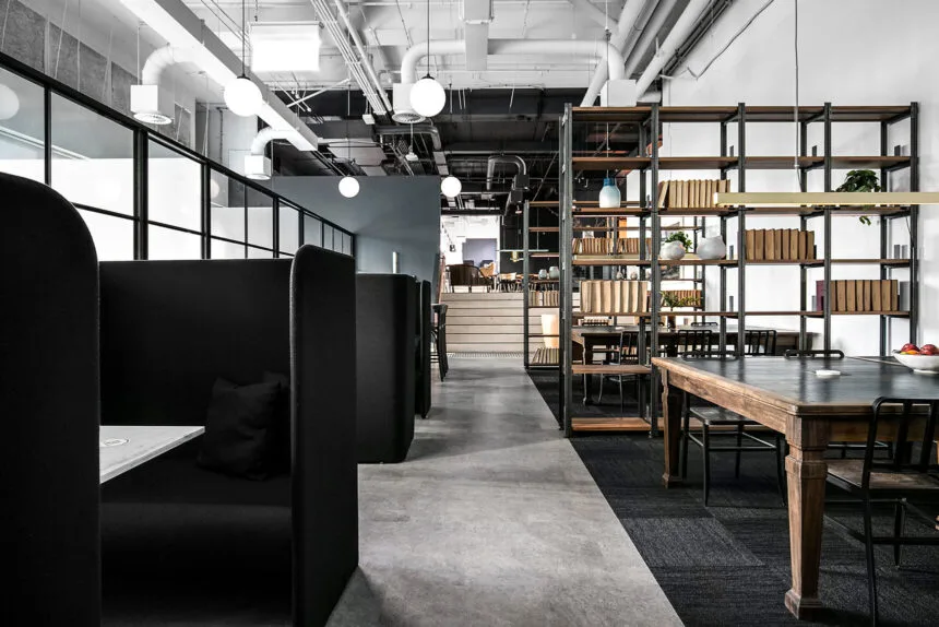 Dark high-backed privacy booths and wooden work tables line a central concrete pathway inside a modern industrial office space featuring open metal shelving, globe pendant lights, and exposed ceiling pipes.