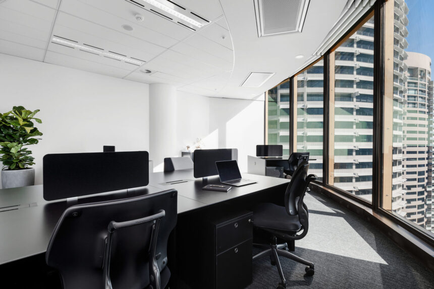 Black modern office desks with ergonomic chairs sit arranged in a bright high-rise office, featuring floor-to-ceiling windows that overlook surrounding city skyscrapers.