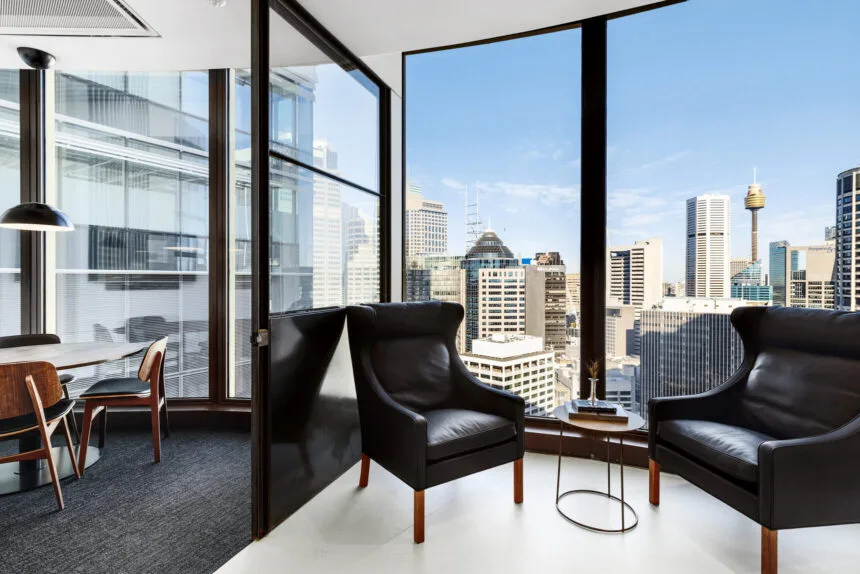 Two black leather armchairs and a small side table sit beside floor-to-ceiling windows inside a modern high-rise office, offering panoramic views of a sunny cityscape skyline.