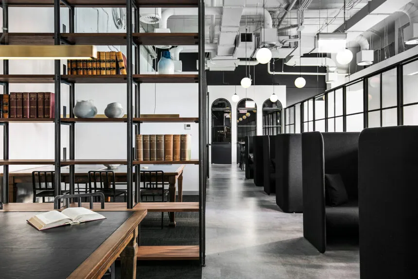 A tall metal shelving unit holding books and decor stands beside high-backed black privacy booths, dividing a modern industrial office space featuring polished concrete floors and exposed ceiling pipes.
