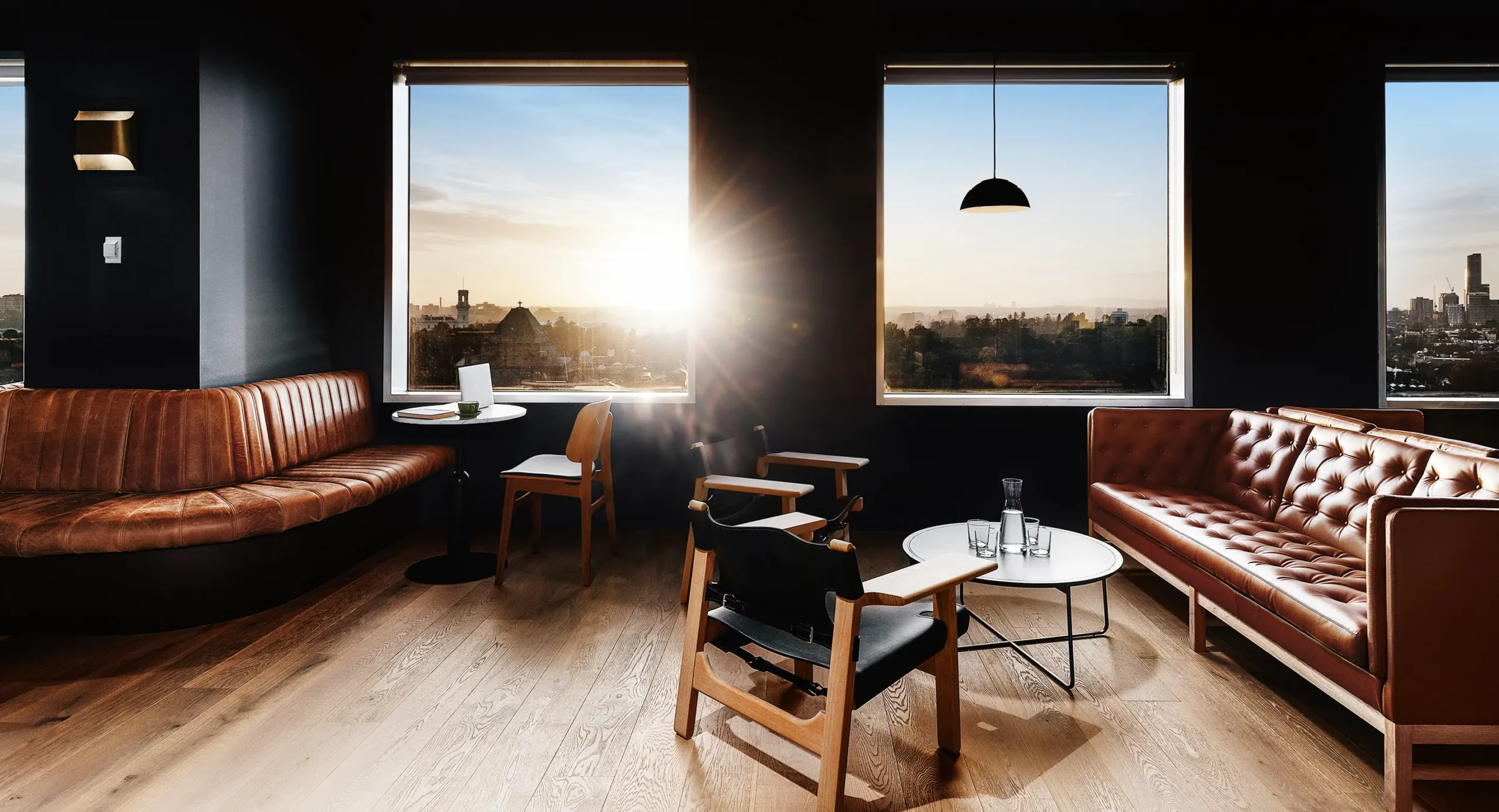Brown leather seating and wooden chairs furnish a dark-walled modern lounge, illuminated by bright sunlight streaming through large windows overlooking a city skyline at sunset.