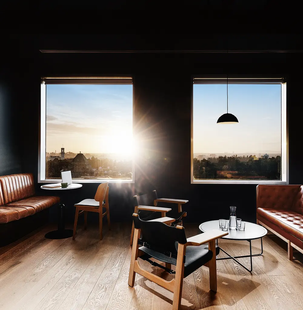Leather seating and wooden armchairs furnish a dark, modern lounge, illuminated by bright sunlight streaming through two large square windows that overlook a city skyline.