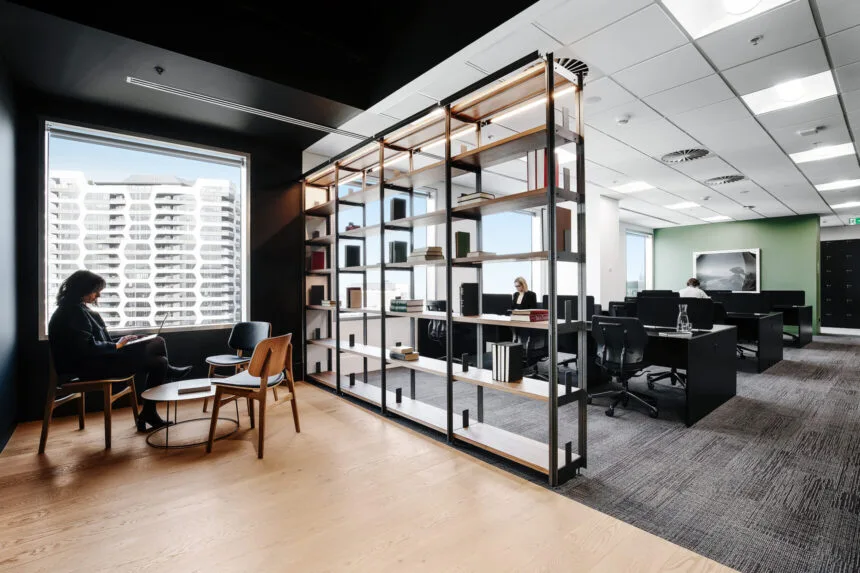 A large metal display shelf divides a cozy modern lounge area, where a person sits near a window, from a bright open office space filled with desks.