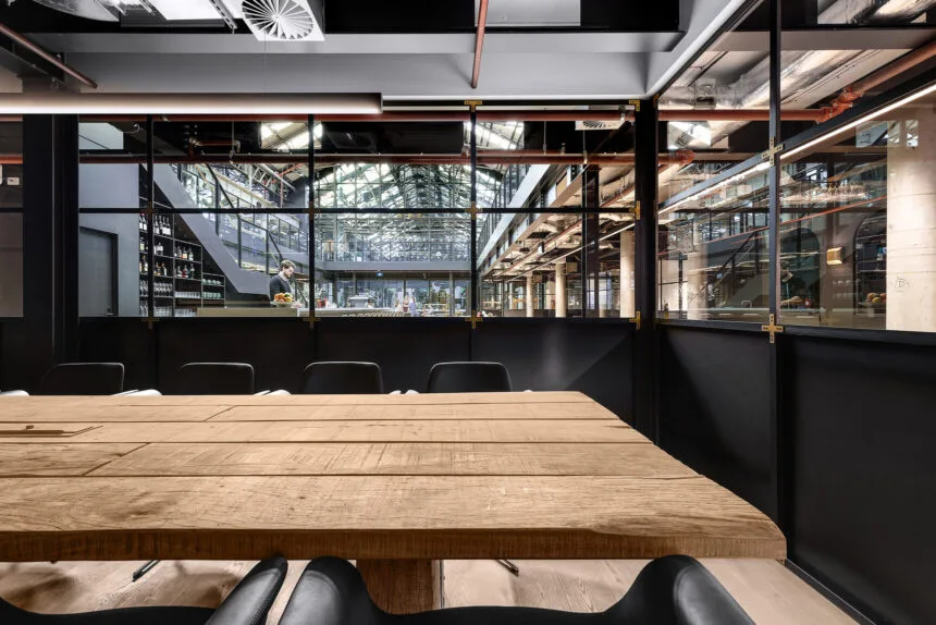 A large, rustic wooden conference table surrounded by black chairs sits inside a glass-partitioned meeting room, overlooking a multi-level, industrial-style indoor atrium filled with exposed pipes and steel beams.