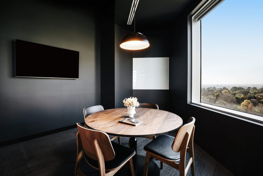 A round wooden meeting table surrounded by four chairs sits beneath a dome pendant lamp inside a modern, black-walled conference room with a large window overlooking treetops.