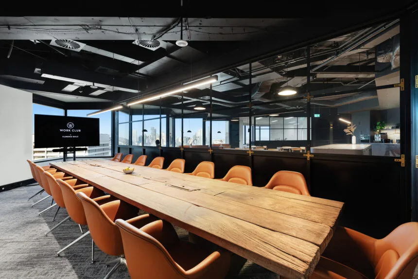 A long rustic wooden conference table lined with cognac leather swivel chairs fills a sleek boardroom with black exposed ceilings and glass walls. A monitor displays: "W WORK CLUB BY FLORENCE GUILD"