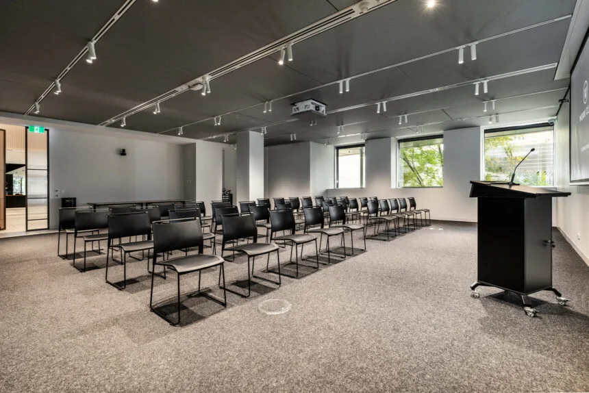 Black chairs arranged in neat rows face a black lectern on wheels inside a modern, carpeted presentation room featuring track lighting on dark ceiling panels and large windows.