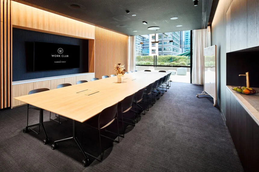 A long light-wood conference table surrounded by dark chairs fills a modern boardroom equipped with a wall-mounted television screen displaying text that reads, "WORK CLUB GLOBAL".