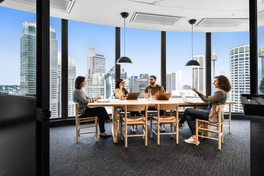 Four colleagues sit around a long wooden table, meeting and working on laptops inside a high-rise office with floor-to-ceiling glass windows offering a sweeping cityscape view.