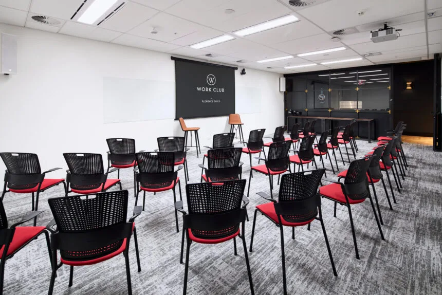 Rows of black and red chairs are arranged facing a projection screen inside a brightly lit presentation room with patterned grey carpet. The screen displays text reading: "W WORK CLUB BY FLORENCE GUILD".