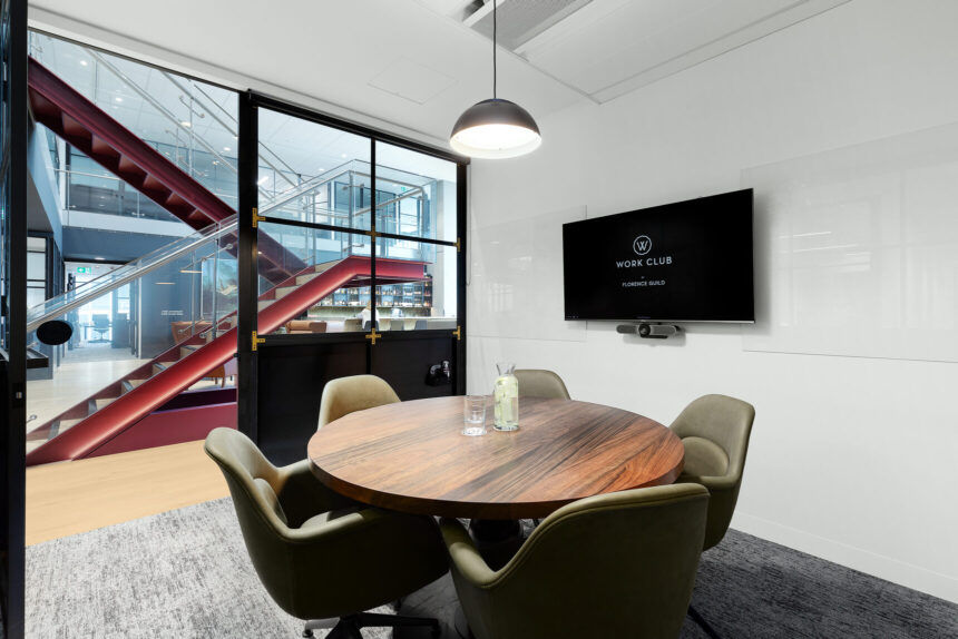 A round wooden table surrounded by green chairs sits in a modern meeting room with a glass wall revealing a red staircase. A wall-mounted TV screen displays: "W WORK CLUB BY FLORENCE GUILD".