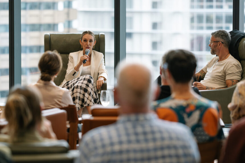A woman in a white blazer speaks into a microphone while seated in a high-backed armchair, addressing an audience inside a modern room with large windows overlooking city skyscrapers.