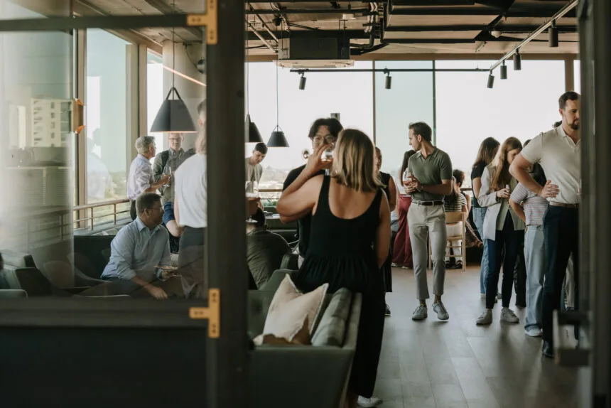 A group of people mingle and converse with drinks in hand inside a modern rooftop lounge featuring floor-to-ceiling glass windows.