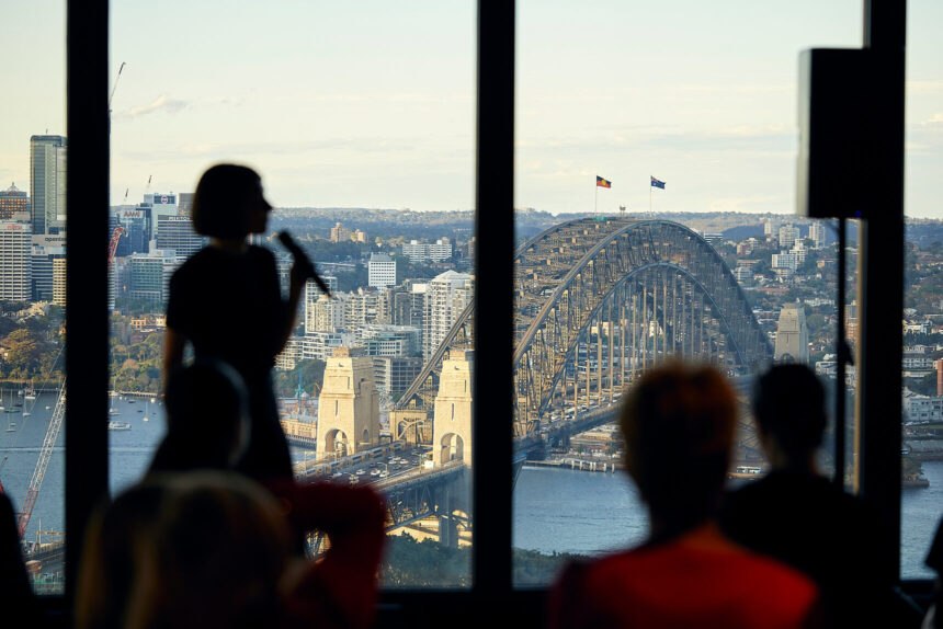 A silhouetted speaker holds a microphone while presenting to an audience in front of large windows overlooking the Sydney Harbour Bridge and the surrounding city skyline.