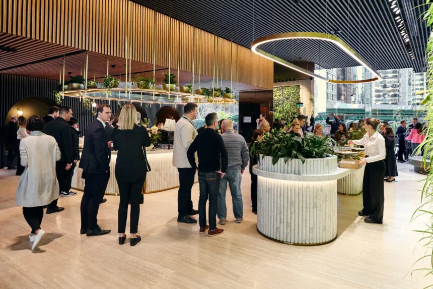People in business attire gather and mingle around modern marble counters at an indoor event, framed by decorative ceiling slats, oval lighting, and potted plants inside a bright, upscale lounge.