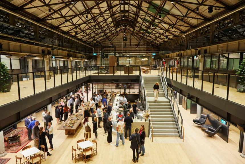 A crowd of people mingles and socializes on the wooden ground floor of a spacious, modern industrial hall featuring mezzanine balconies, a central staircase, and exposed steel roof trusses.