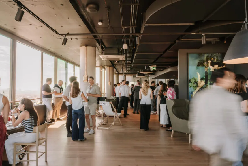 A crowd of people mingles and converses inside a modern high-rise venue featuring wooden flooring, dark industrial ceilings, and large floor-to-ceiling windows overlooking a skyline. Text on a sling chair reads: AIX.