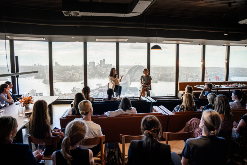 Two speakers present to a seated audience inside a high-rise room featuring expansive floor-to-ceiling windows with a panoramic view of a city harbor and bridge.