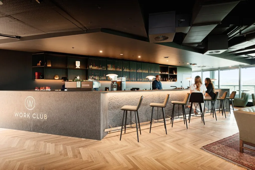 A grey stone bar counter featuring the text "WORK CLUB" hosts a bartender and seated patrons inside a modern lounge with dark ceilings and herringbone wooden flooring.