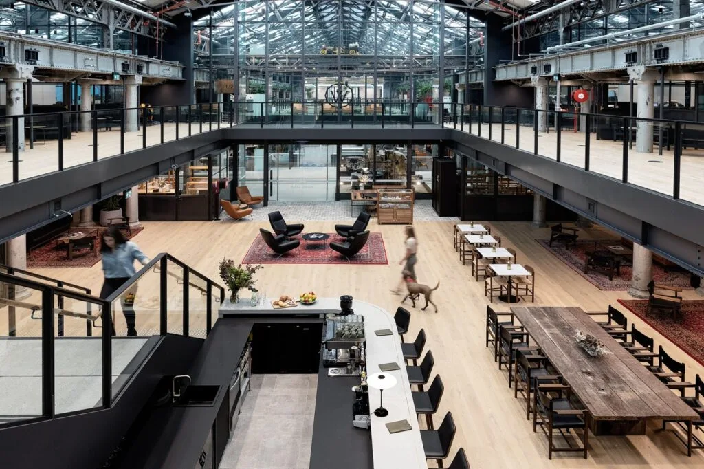 People walk through a spacious, open-plan industrial atrium featuring a long bar counter, lounge chairs, and dining areas beneath a high glass roof and surrounding mezzanine level.