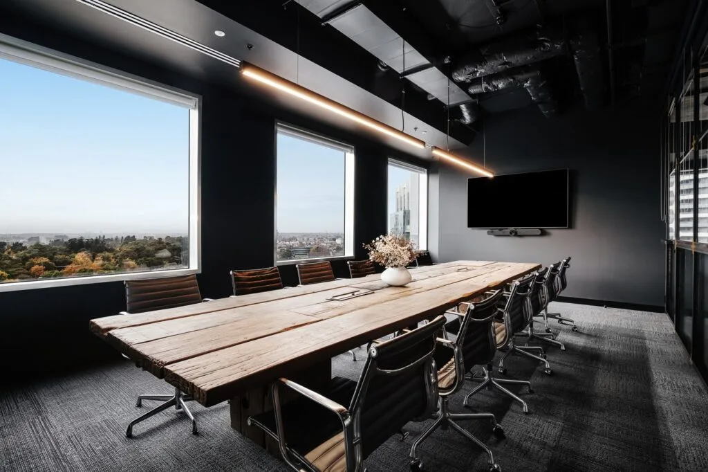 A long wooden conference table surrounded by black executive chairs sits in a sleek boardroom featuring large windows showing a city view, dark walls, overhead lights, and a wall-mounted monitor.