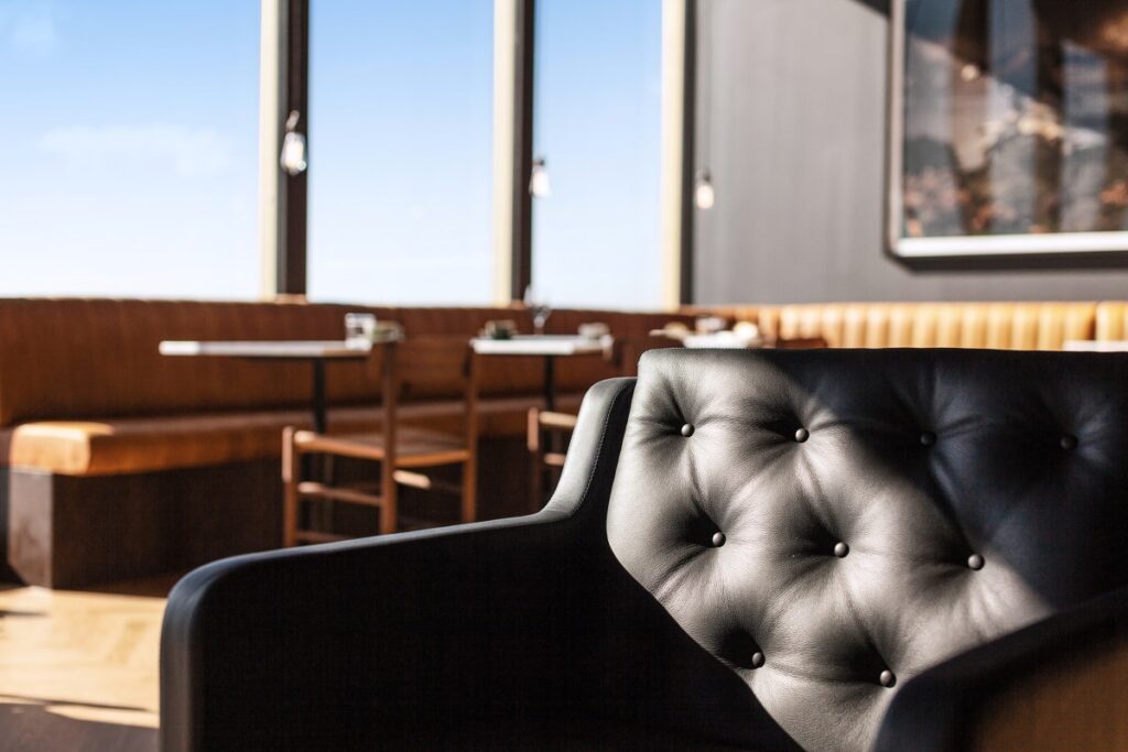 A black tufted leather armchair sits in the foreground of a sunlit restaurant interior featuring floor-to-ceiling windows, tan leather booth seating, and small wooden dining tables.