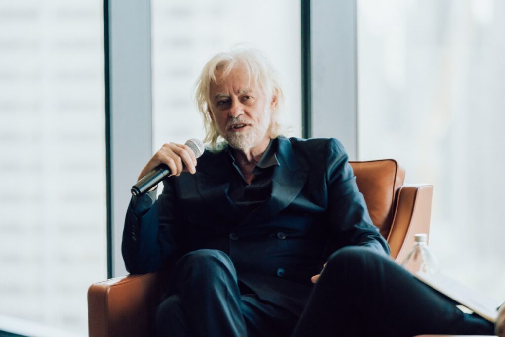 A white-haired man in a dark suit sits in a brown leather armchair, speaking into a microphone in front of large floor-to-ceiling windows.