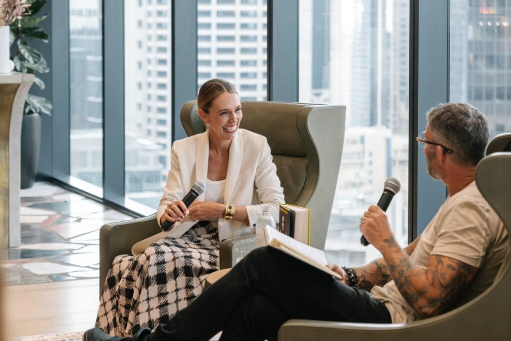 A smiling woman in a white blazer and a tattooed man sit in armchairs holding microphones, conversing during an event in a high-rise room with floor-to-ceiling city views.