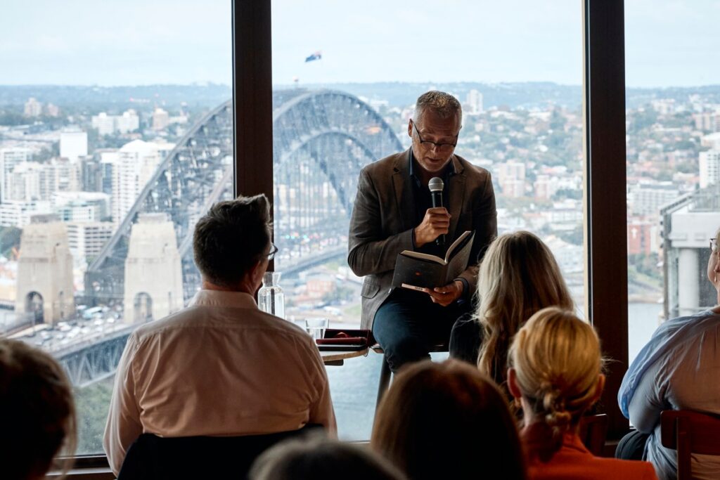 A man in a jacket reads from an open book into a microphone before a seated audience, positioned in front of large windows overlooking the Sydney Harbour Bridge.