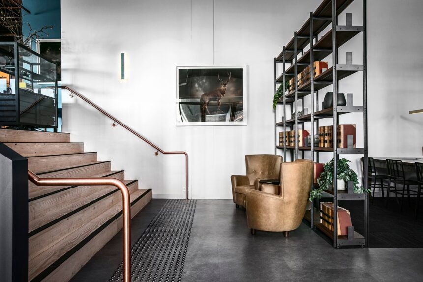 Two tan leather armchairs rest beside a tall metal bookcase in a modern lobby, next to wooden stairs with copper handrails and a framed stag picture on the wall.