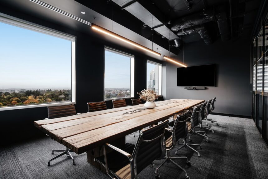 A long rustic wooden conference table surrounded by black leather chairs extends through a modern boardroom featuring large city-view windows, overhead linear pendant lights, and dark walls.