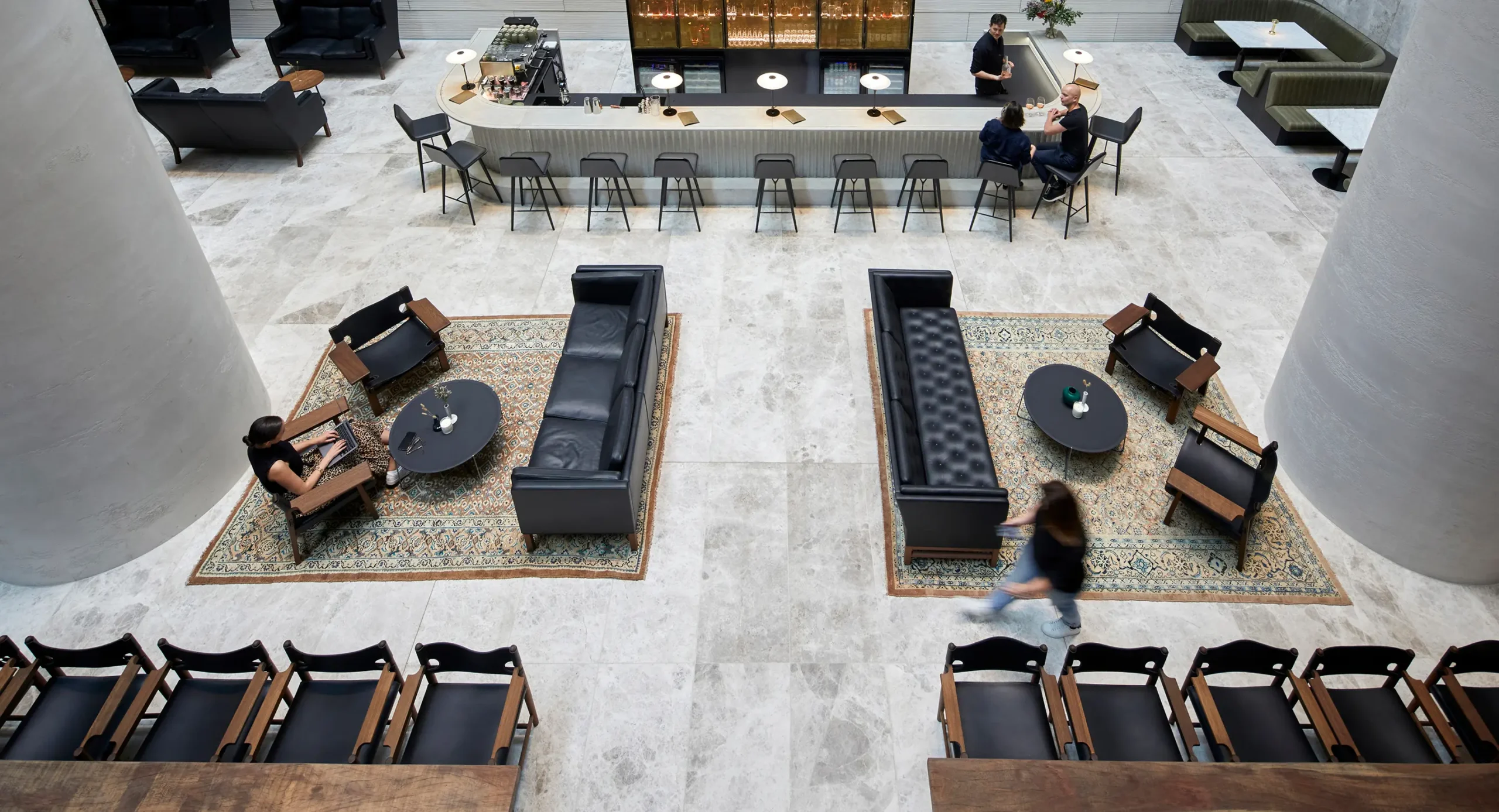 Patrons sit and walk through a modern lounge, conversing at a long bar and working in leather seating areas set on patterned rugs across a wide marble floor.