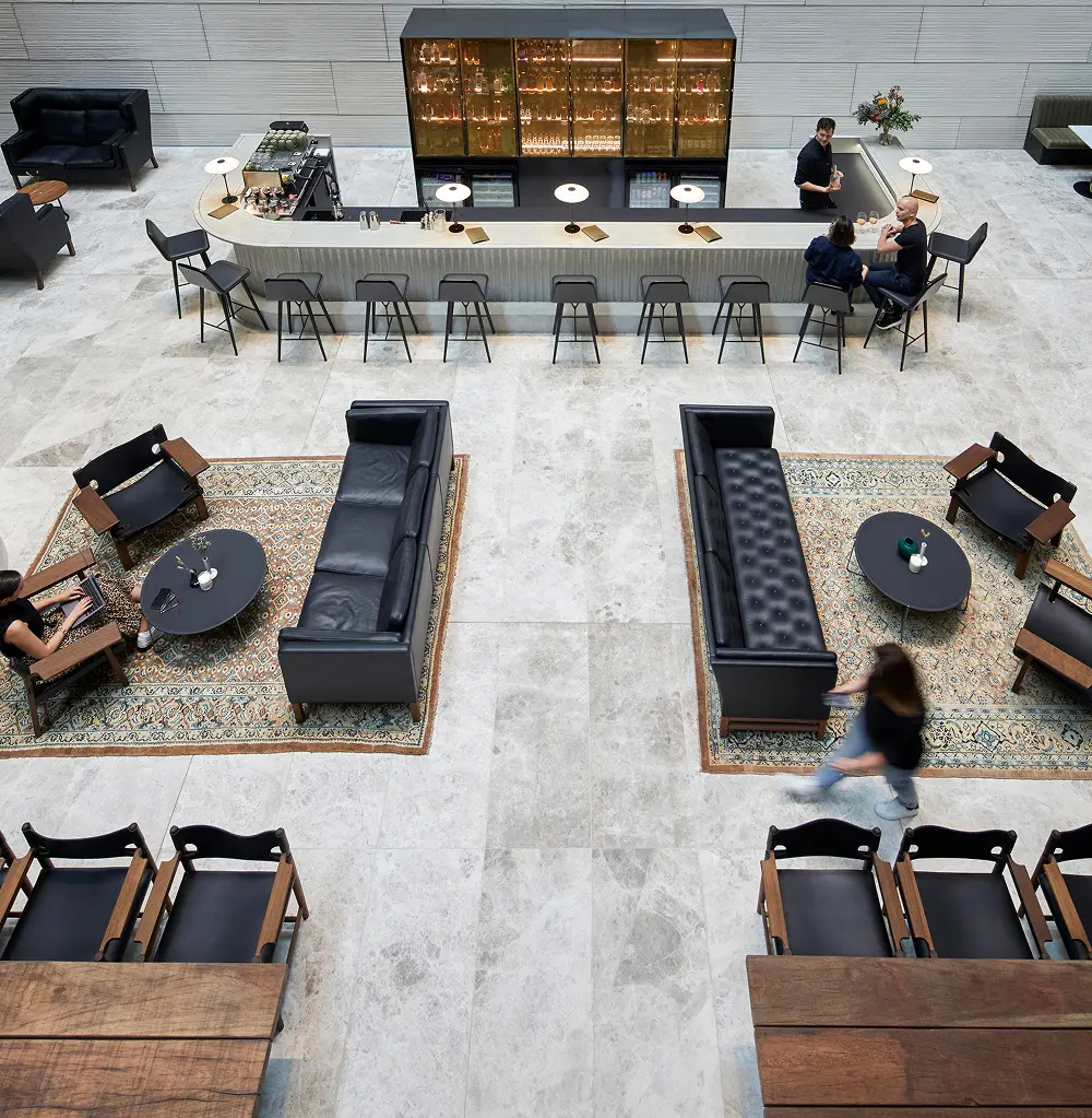 A high-angle view shows people working, walking, and socializing around a central bar counter and black leather seating arrangements on patterned rugs in a spacious, modern tiled lobby.
