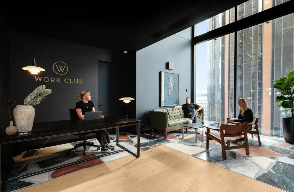 A receptionist sits at a dark desk while two people relax in an adjacent lounge beside floor-to-ceiling windows inside a modern office. Gold wall text reads, "W WORK CLUB".
