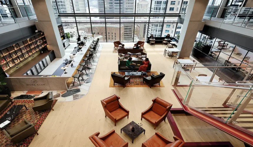 People converse across various seating areas in a high-ceilinged modern lounge featuring leather armchairs, a bar, and floor-to-ceiling windows overlooking city skyscrapers. Text on the bar reads: “W WORK CLUB”.
